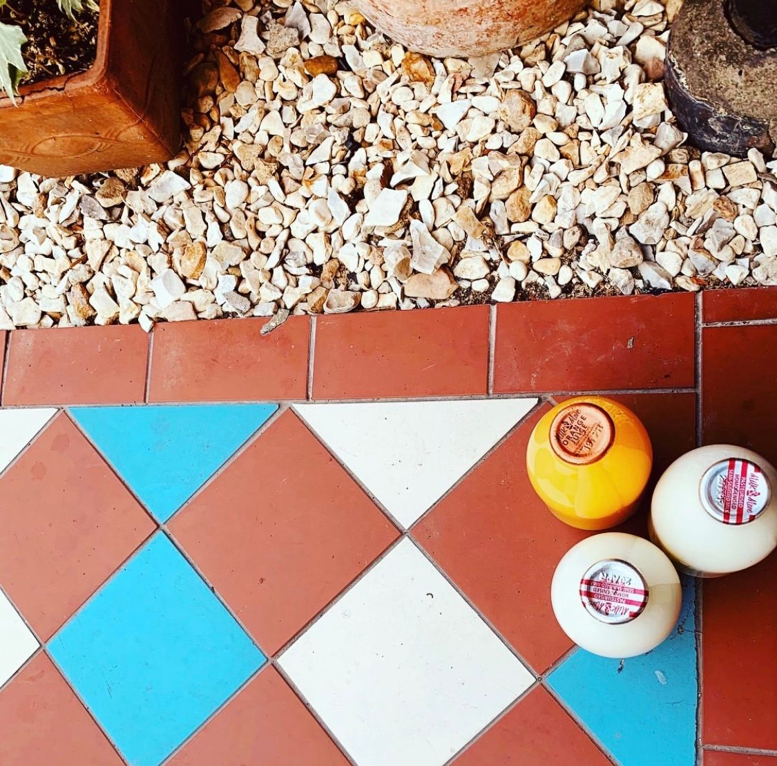 Three glass bottles of milk, one orange and two white, sit on a patterned terracotta tile floor beside a gravel bed.