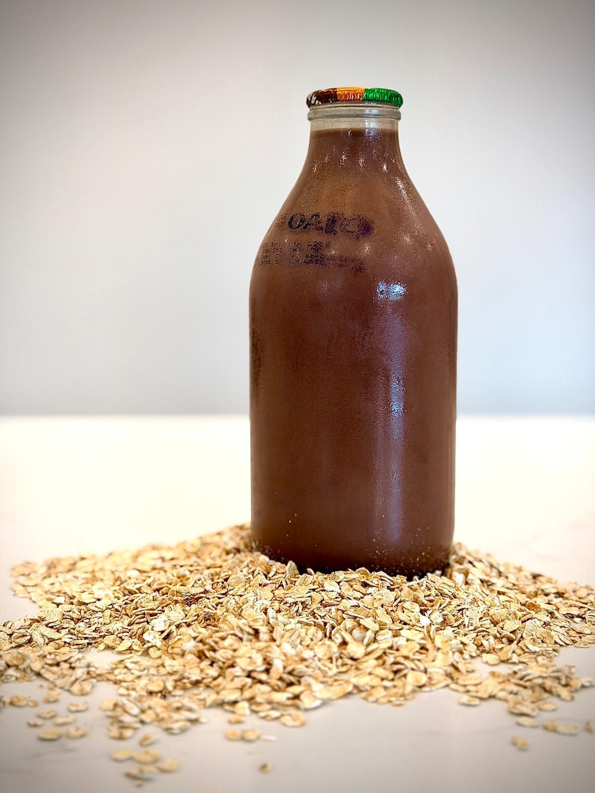 A bottle of brown, chocolate-colored oat milk sitting on a pile of raw, light-colored rolled oats on a white surface.