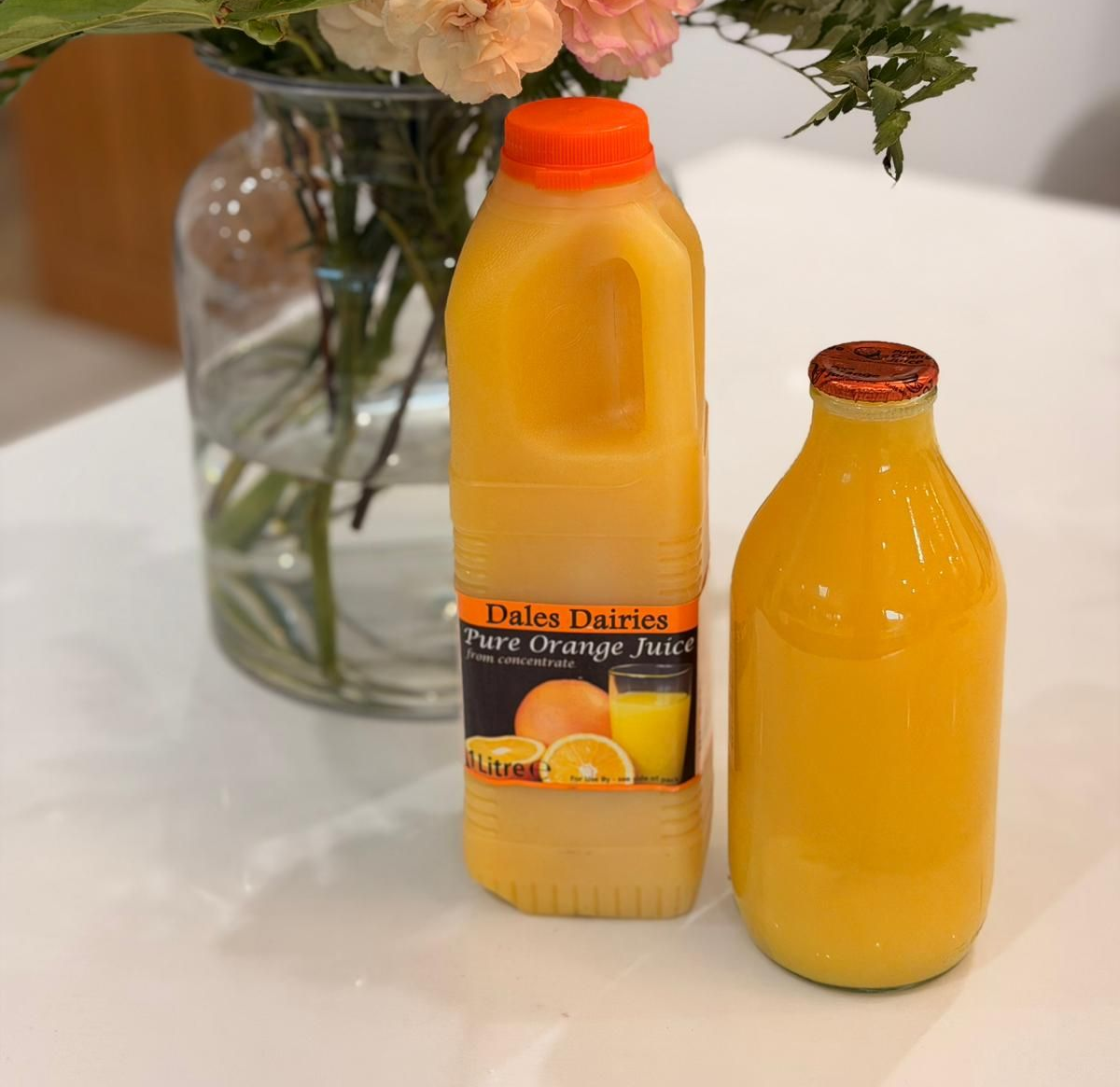 Two bottles of orange juice stand on a white table next to a glass vase with flowers.
