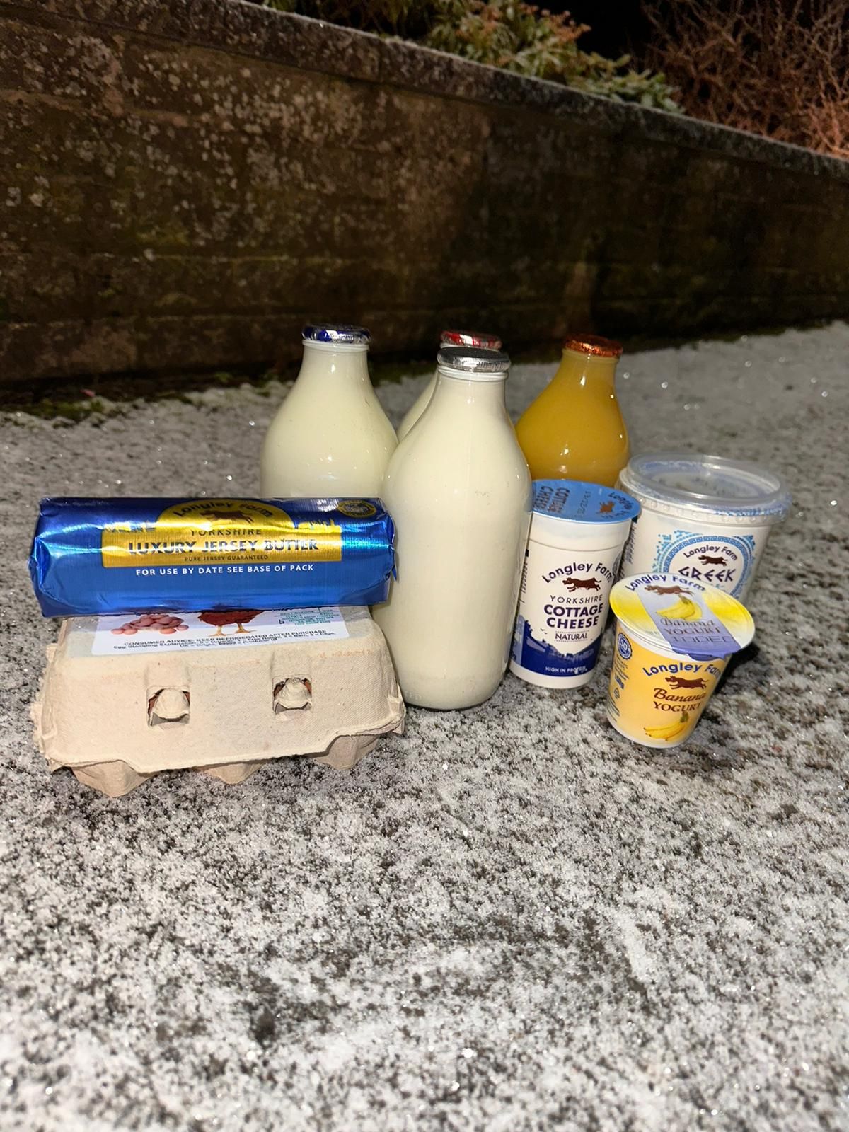 A doorstep delivery of milk in glass bottles, a carton of eggs, butter, yogurt, and orange juice on a snowy surface.