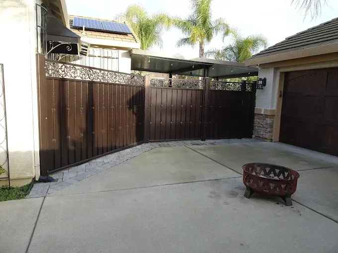 Brown gate with decorative top, leading to a driveway and garage, and a fire pit.