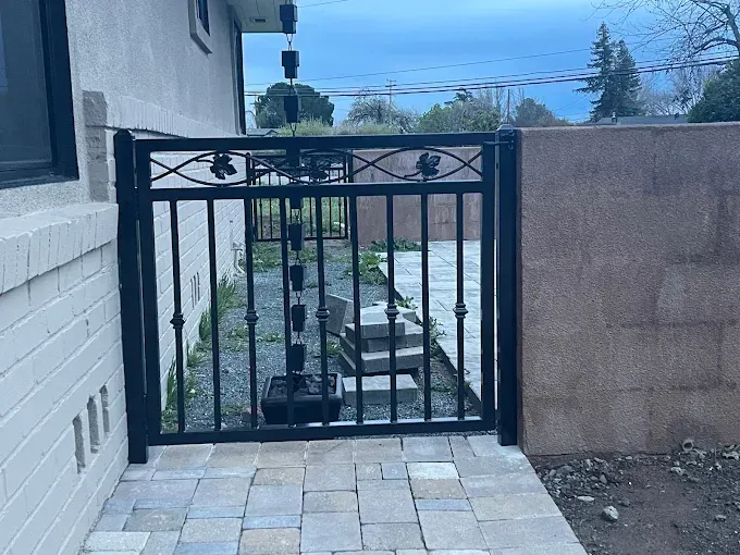 Black metal gate between a brick house and a stone wall, leading to a path with a rain chain.