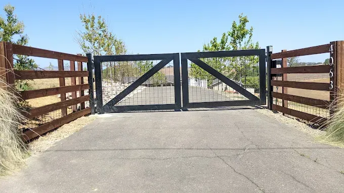 Black double gate with metal mesh and wooden fence, leading to a driveway.