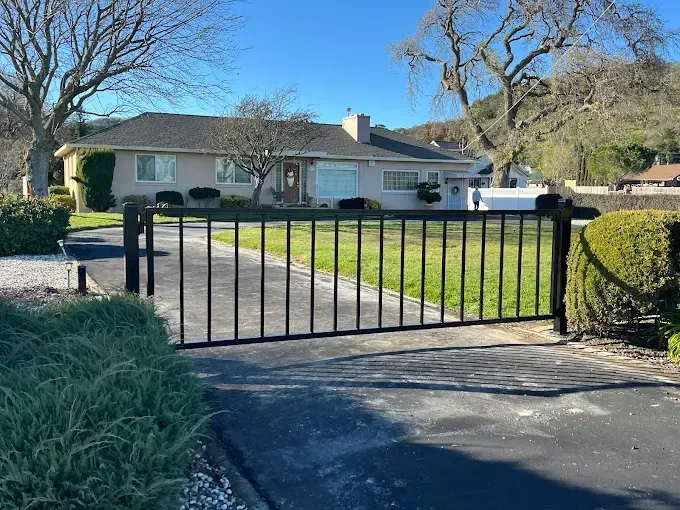 Black metal gate in front of a light-colored ranch-style home, on a sunny day.