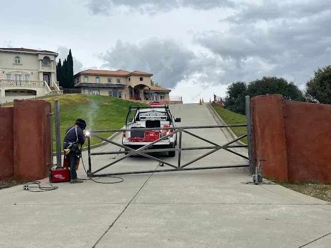 A person welding a metal gate in front of two large houses on a hillside; a truck is behind the gate.