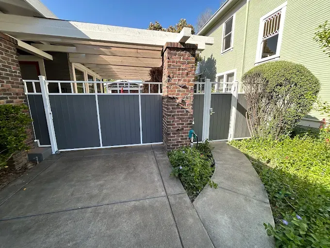 Gray gates open onto a concrete driveway, flanked by a brick pillar and a green house.