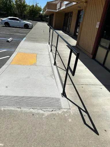 Sidewalk with ramp and black handrail in front of shops; white car parked in background.