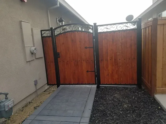Wooden gate with metal frame and decorative scrollwork. Gray walkway, brown mulch, beige wall.