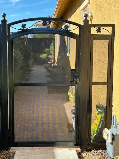 Black metal gate with arched top and screen door, leading to a path in a yard.