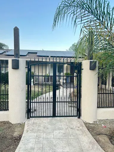 Black metal gate in front of a house, set between two cream-colored pillars with exterior lighting and a concrete walkway.
