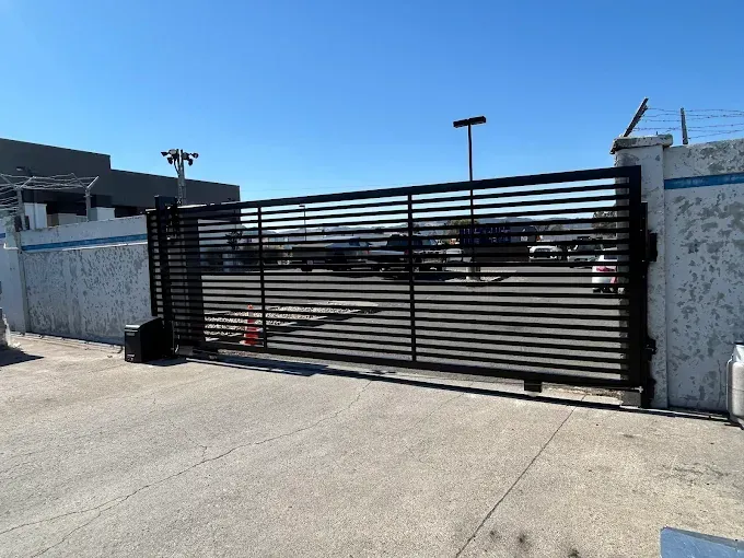 Black metal sliding gate, open, in concrete wall with barbed wire, leading to a parking area.