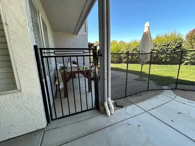 Black gate on a patio, leading to a yard with a fence and umbrella. Clear sky.