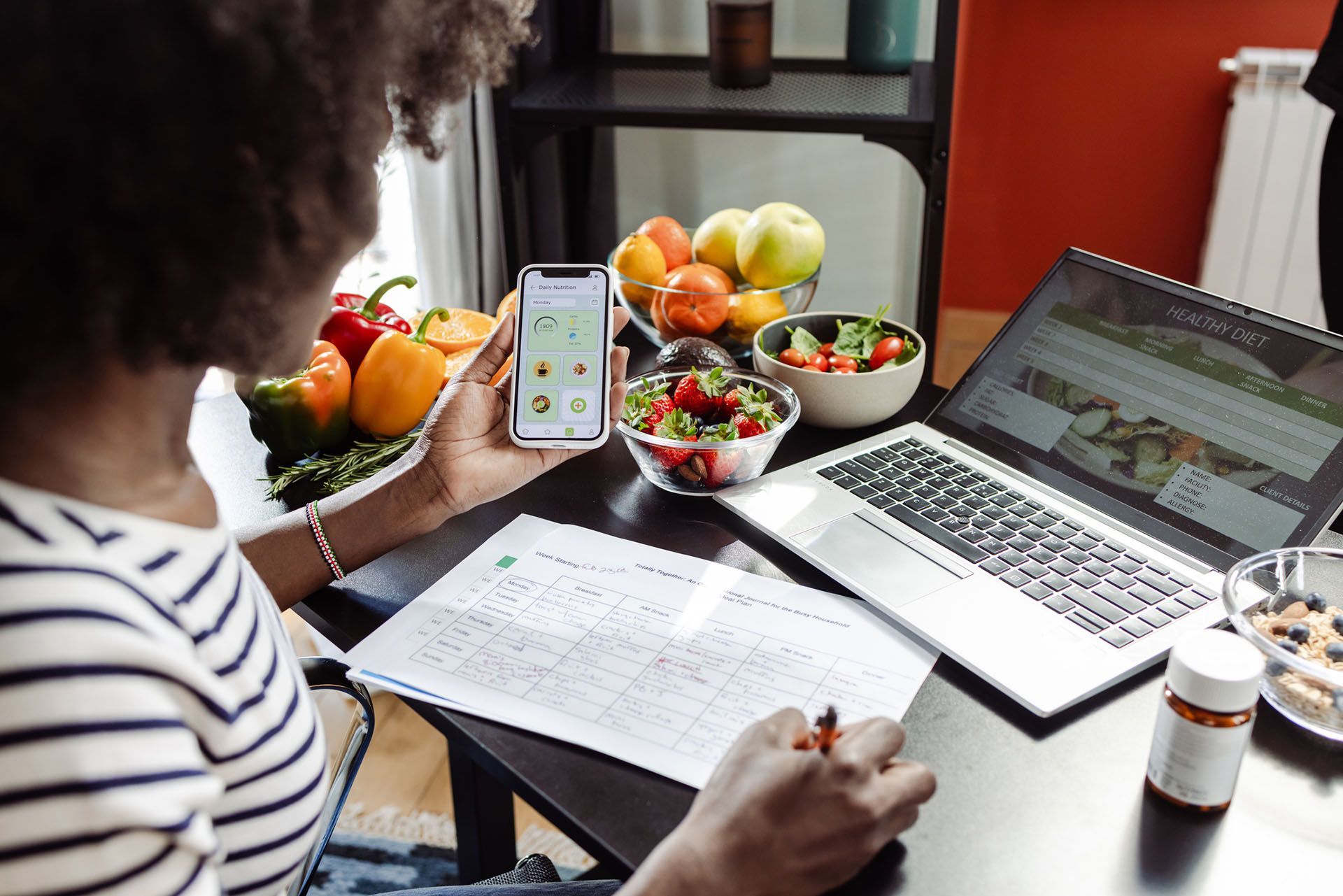 A woman at a desk with a laptop, phone, food, and planning a healthy meal