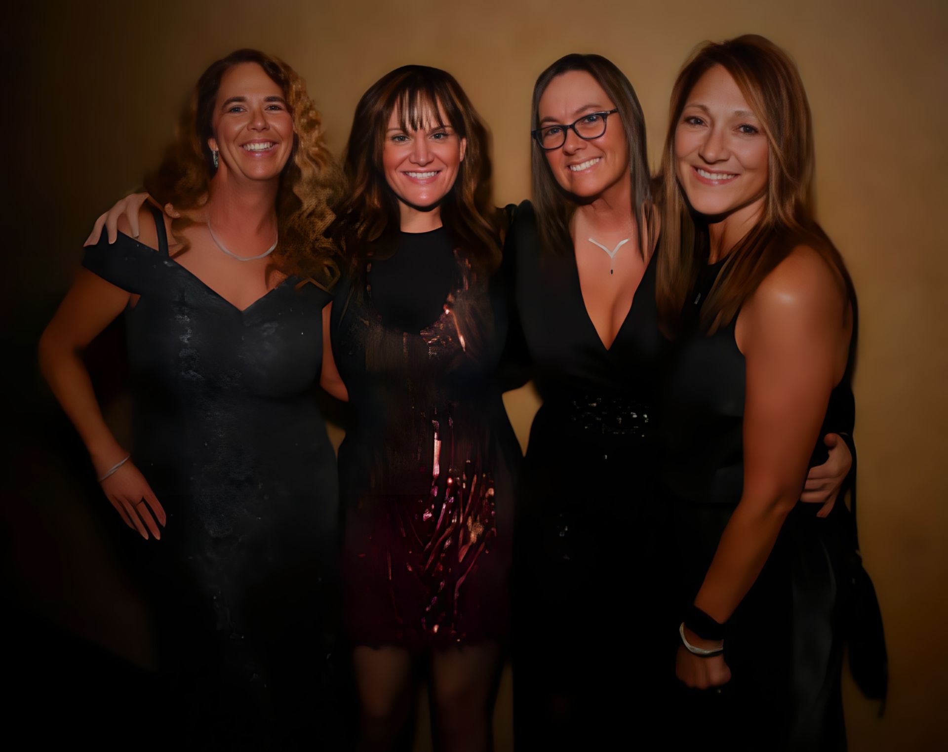 Four women in elegant dark dresses pose side-by-side and smile at the camera in a dimly lit, indoor setting.