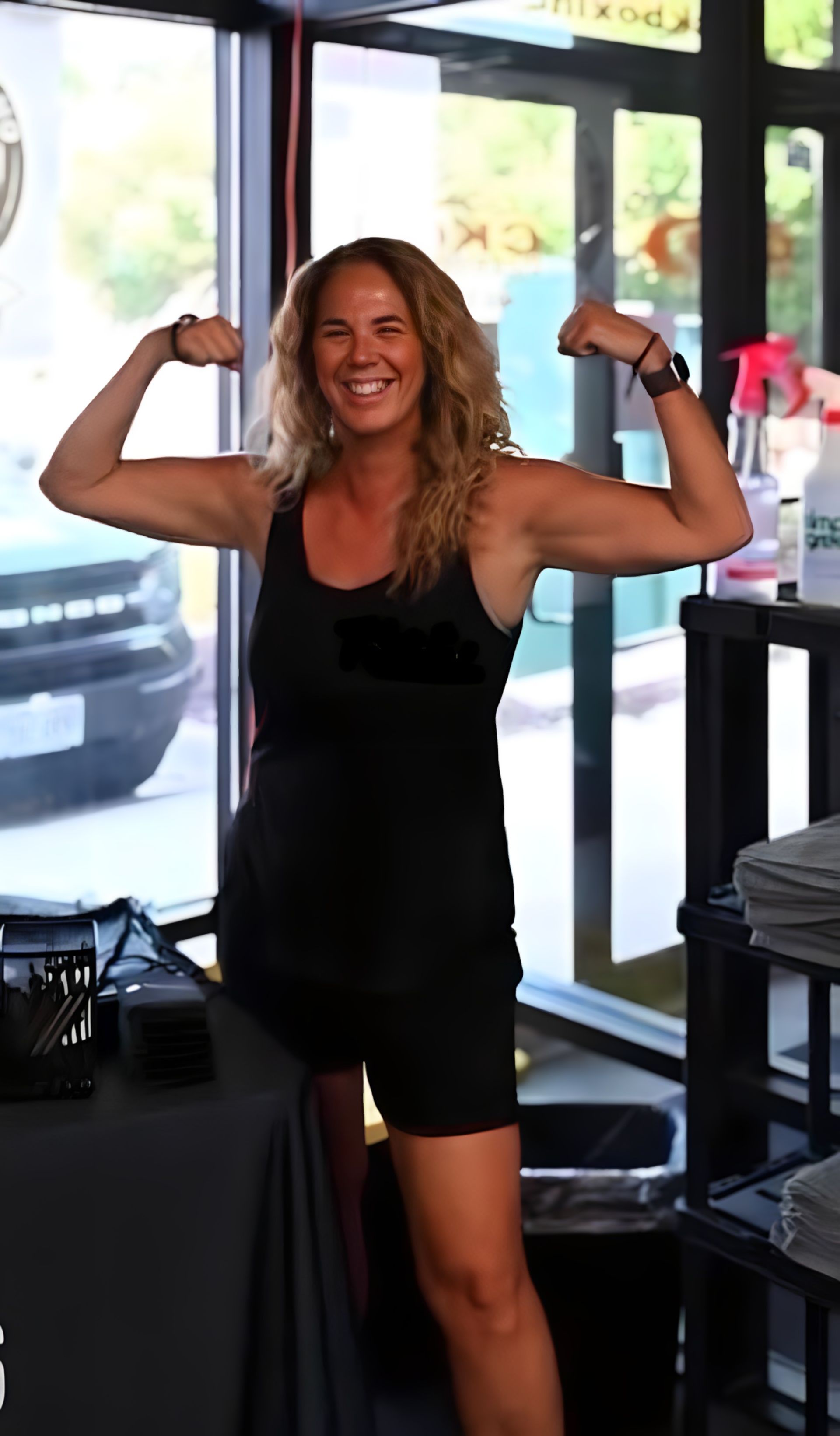 A smiling person wearing a black tank top and shorts flexes their arms in a gym setting near a window.