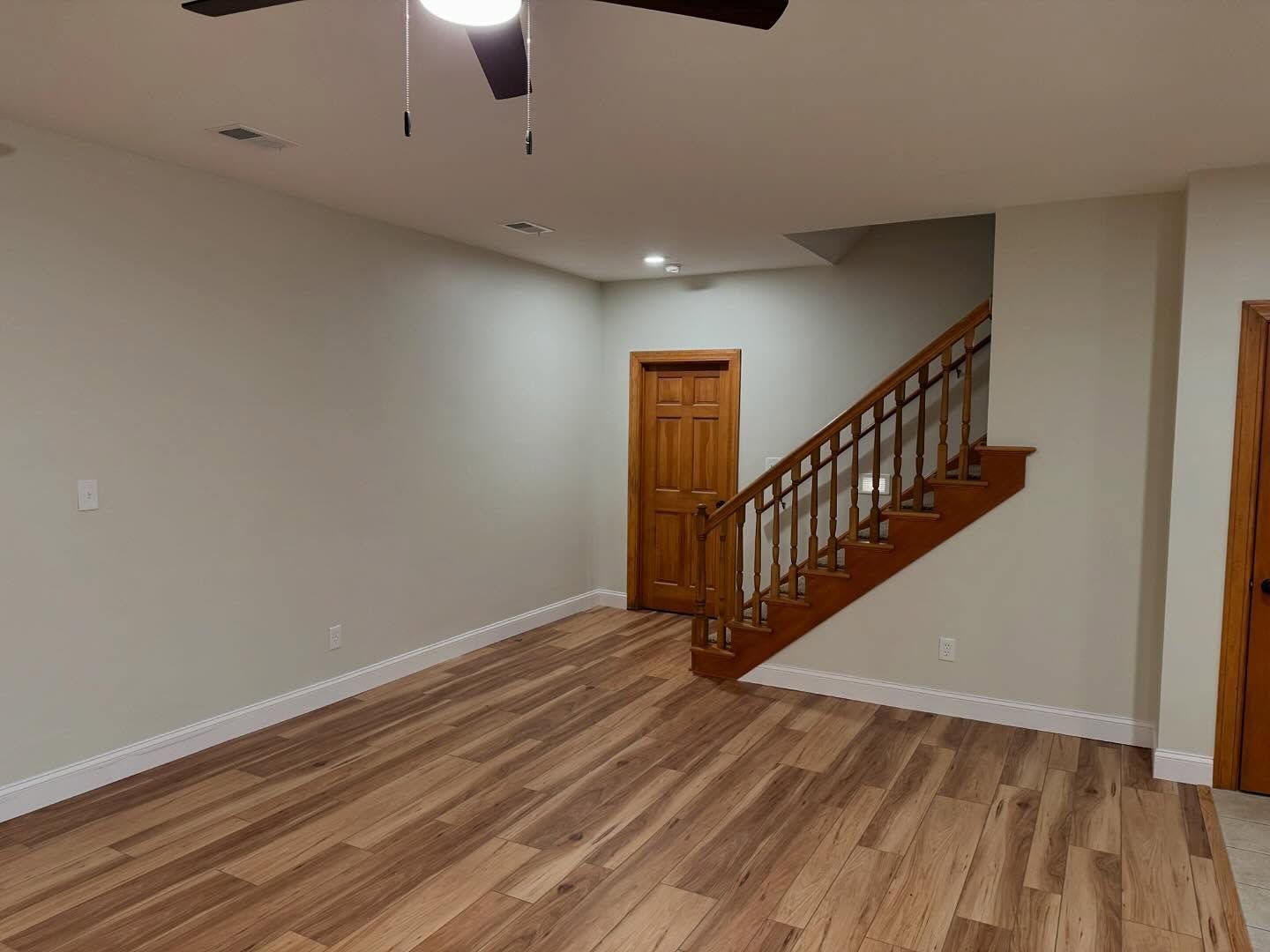 Empty living room with wood floor, beige walls, and a staircase beside a wooden door.