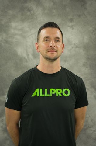 Man standing against a gray backdrop, wearing a black ALLPRO T-shirt with green lettering. Man standing against a gray backdrop, wearing a black ALLPRO T-shirt with green lettering.