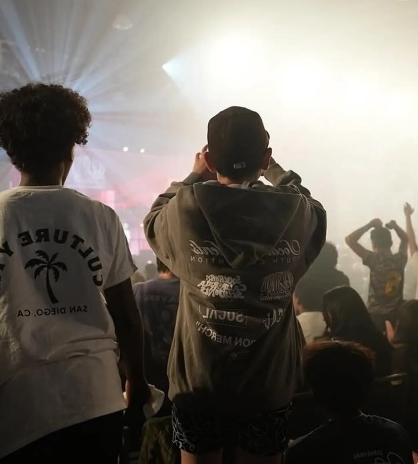 Two people in a crowd with bright lights, one wearing a shirt with a palm tree, the other with a hoodie; concert setting.