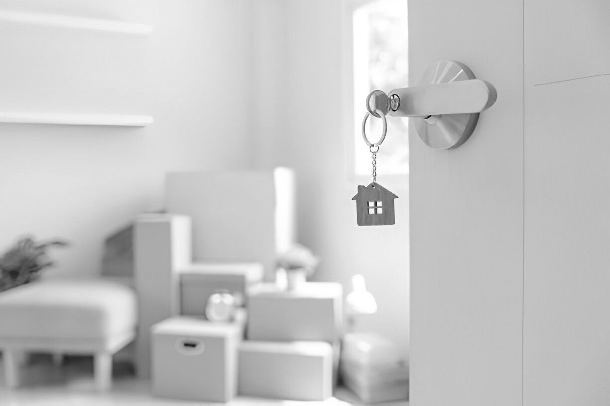 Door with keys and house keychain opens to a room with stacked moving boxes, in grayscale.