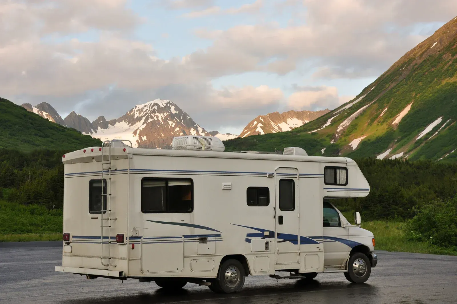 A white motorhome parked on an asphalt lot in front of mountains with snow-covered peaks under a partly cloudy sky.