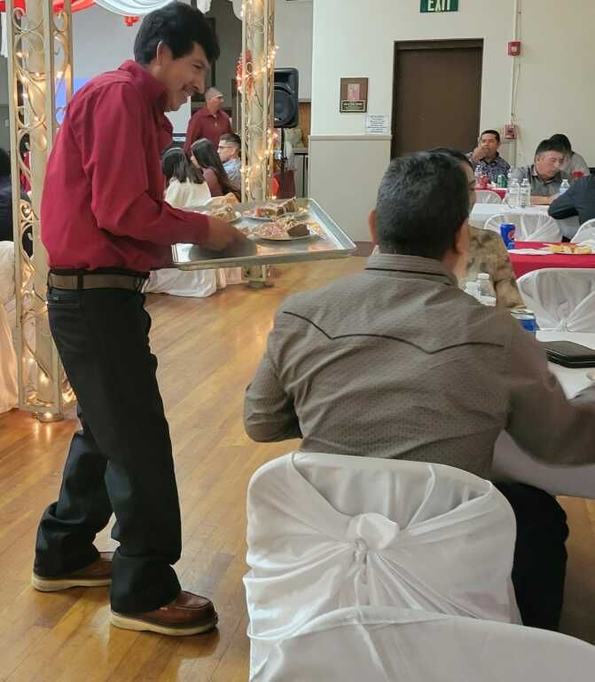 A waiter in a red shirt serves food on a large tray at a busy event with decorated tables and guests dining and conversing.