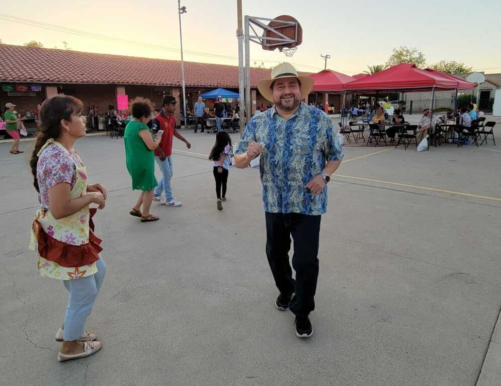 A man in a hat and floral shirt joyfully dances in an outdoor gathering. 