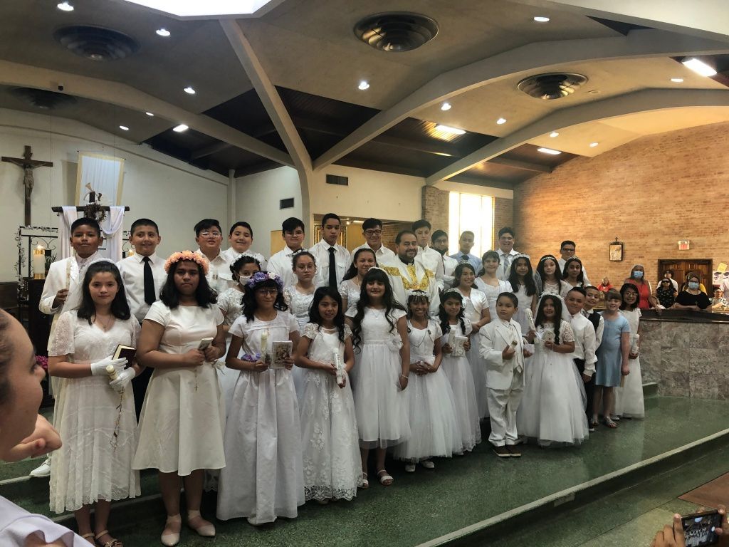 A group of children in white dresses and suits stand on steps inside a church, celebrating a religious ceremony.