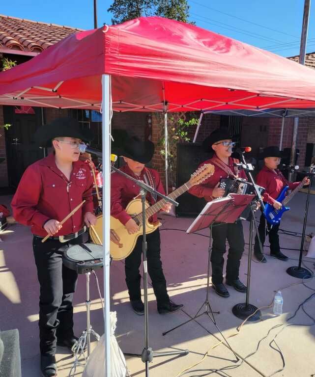 A band of four musicians performs under a red canopy, wearing matching red shirts and black cowboy hats.