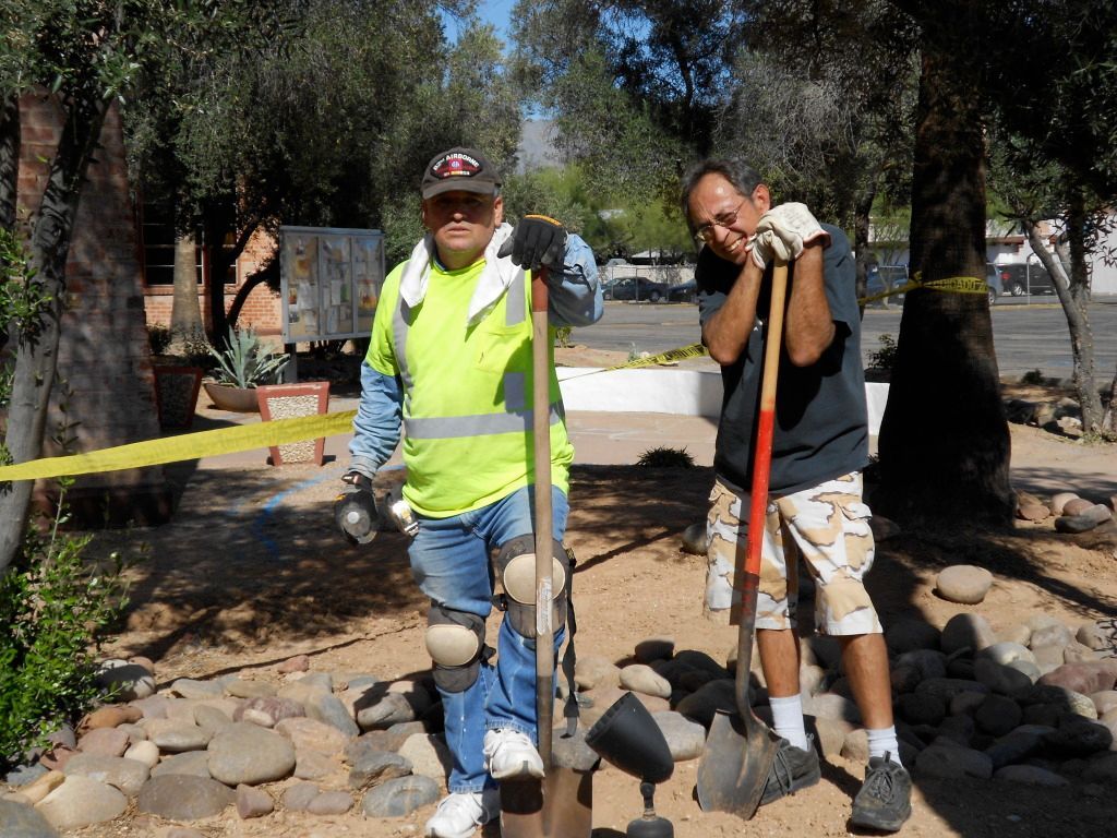Two men are resting on their shovels in a sunny garden area. 