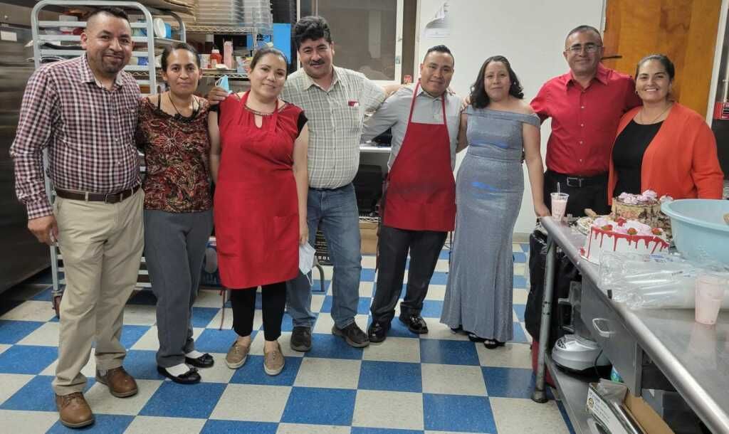 A group of eight people stands in a kitchen smiling, some in uniforms. 
