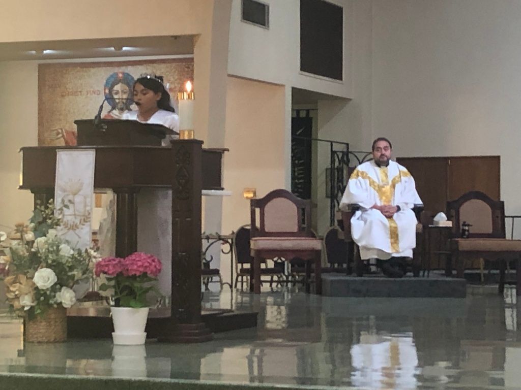 A young girl reads at a church lectern adorned with a candle and flowers.
