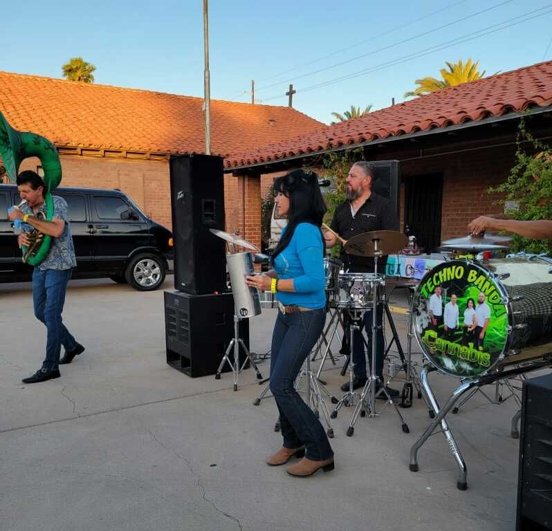 Outdoor band performance with a woman in blue holding a percussion instrument, a drummer, and a saxophonist.