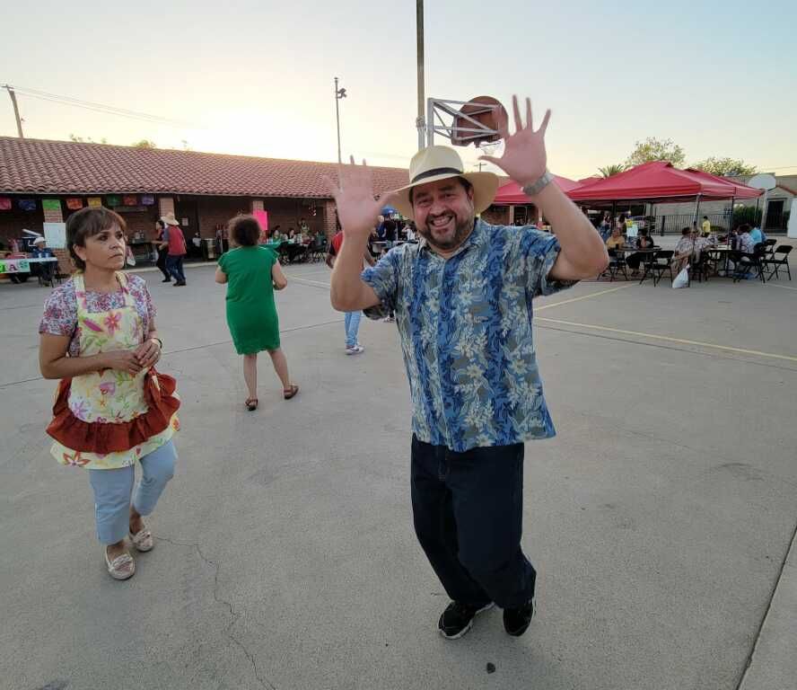 A man in a straw hat and floral shirt joyfully raises his hands at an outdoor gathering. 