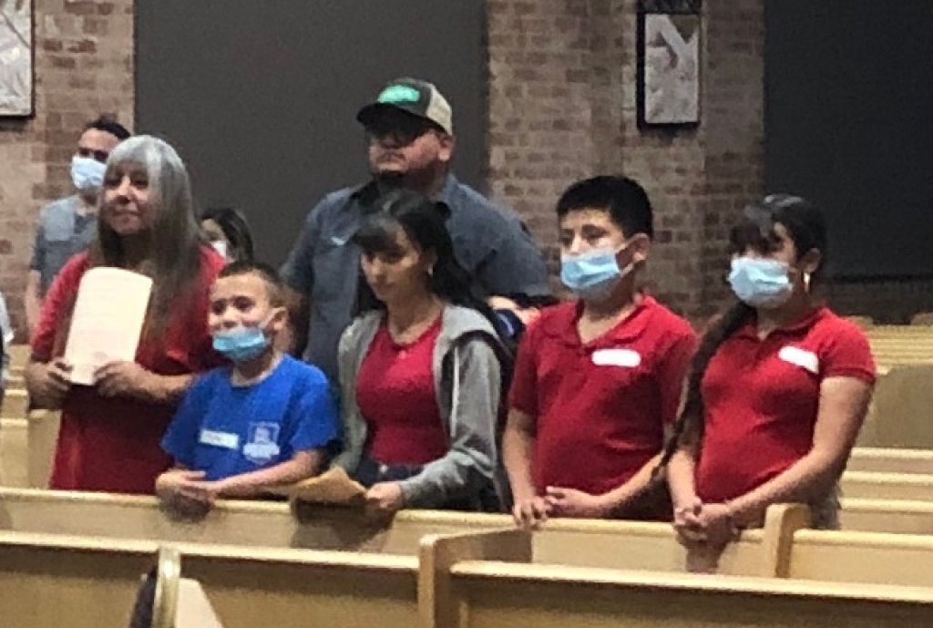 A group of people, including children and adults, stands in church pews.