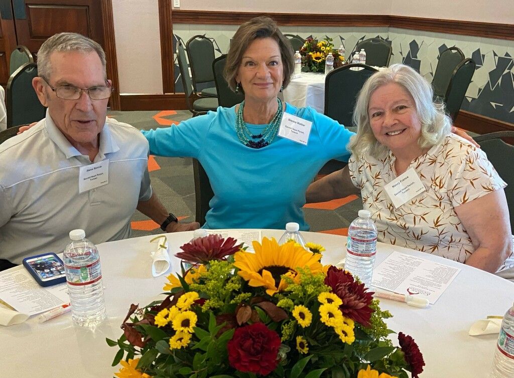 Three people sit at a round table with a floral centerpiece of sunflowers and roses, smiling warmly.