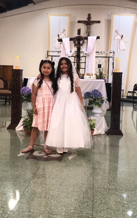 Two young girls stand smiling in front of a decorated altar with white drapes and flowers in a church. 