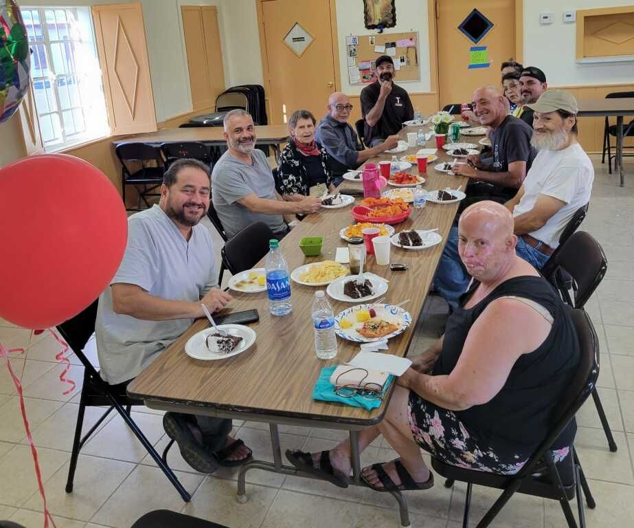 A group of people are smiling and enjoying a meal around a long table, decorated with plates of food and colorful napkins.
