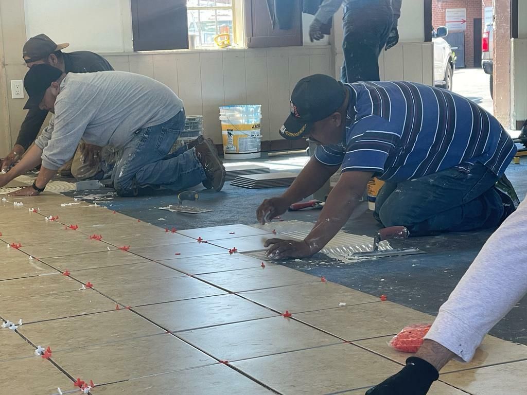 Three workers in casual clothing are on their knees, laying tiles on a floor. 