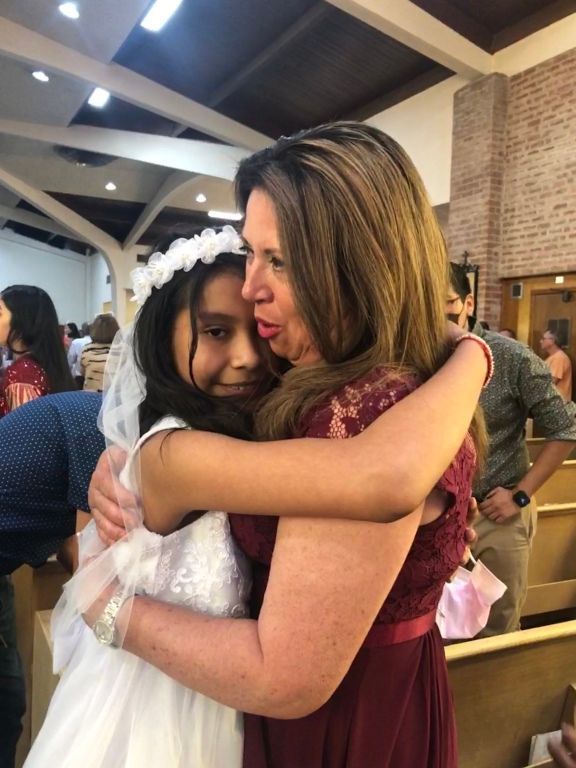 A woman in a burgundy dress embraces a young girl in a white dress and veil inside a church.
