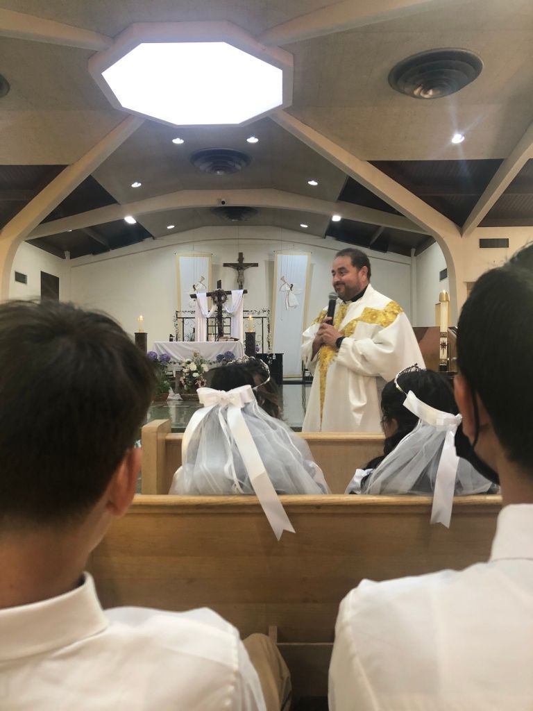 A priest in white and gold robes speaks into a microphone inside a church.