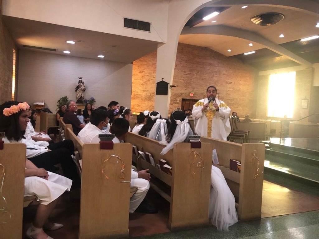 A priest in white and gold robes speaks to children dressed in white for a special occasion.