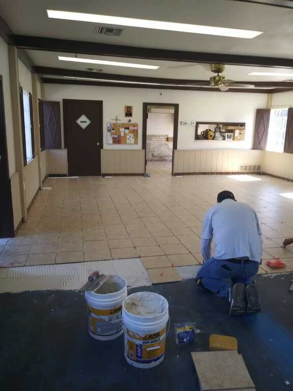 A worker in a blue shirt kneels on a tiled floor, applying tiles with adhesive. 