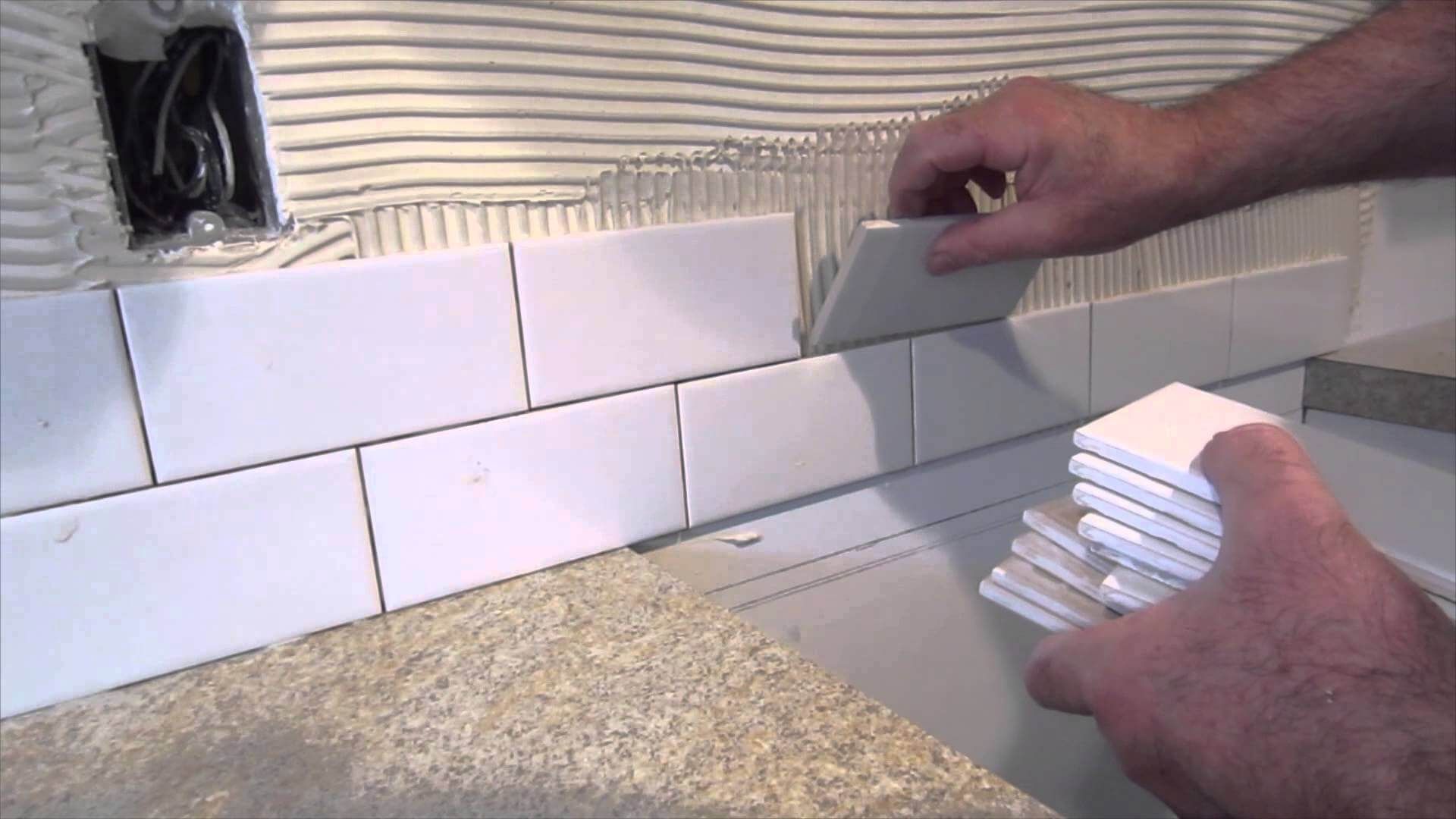 Hands installing white subway tiles on a kitchen backsplash above a countertop