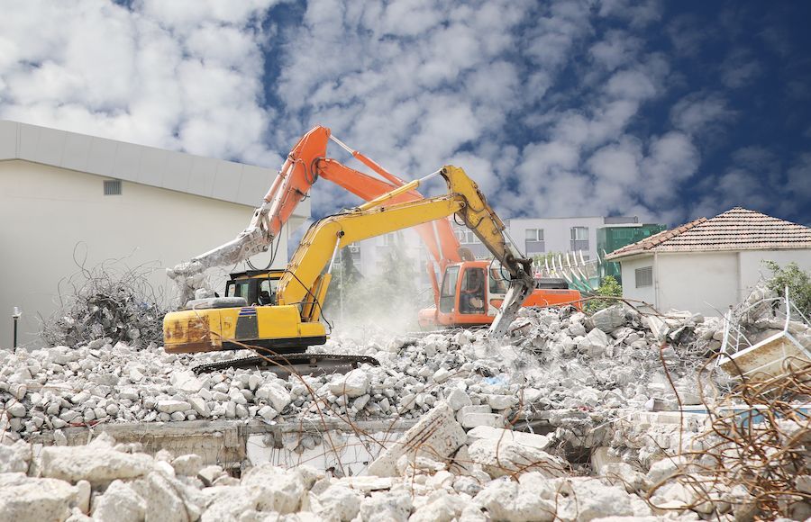 Excavator demolishing a rubble pile beside a white building under a cloudy sky