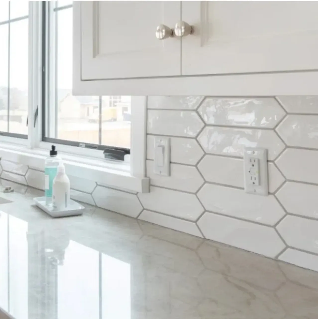 Bright white kitchen countertop with hexagon tile backsplash, window, cabinet, soap dispenser, and outlet.