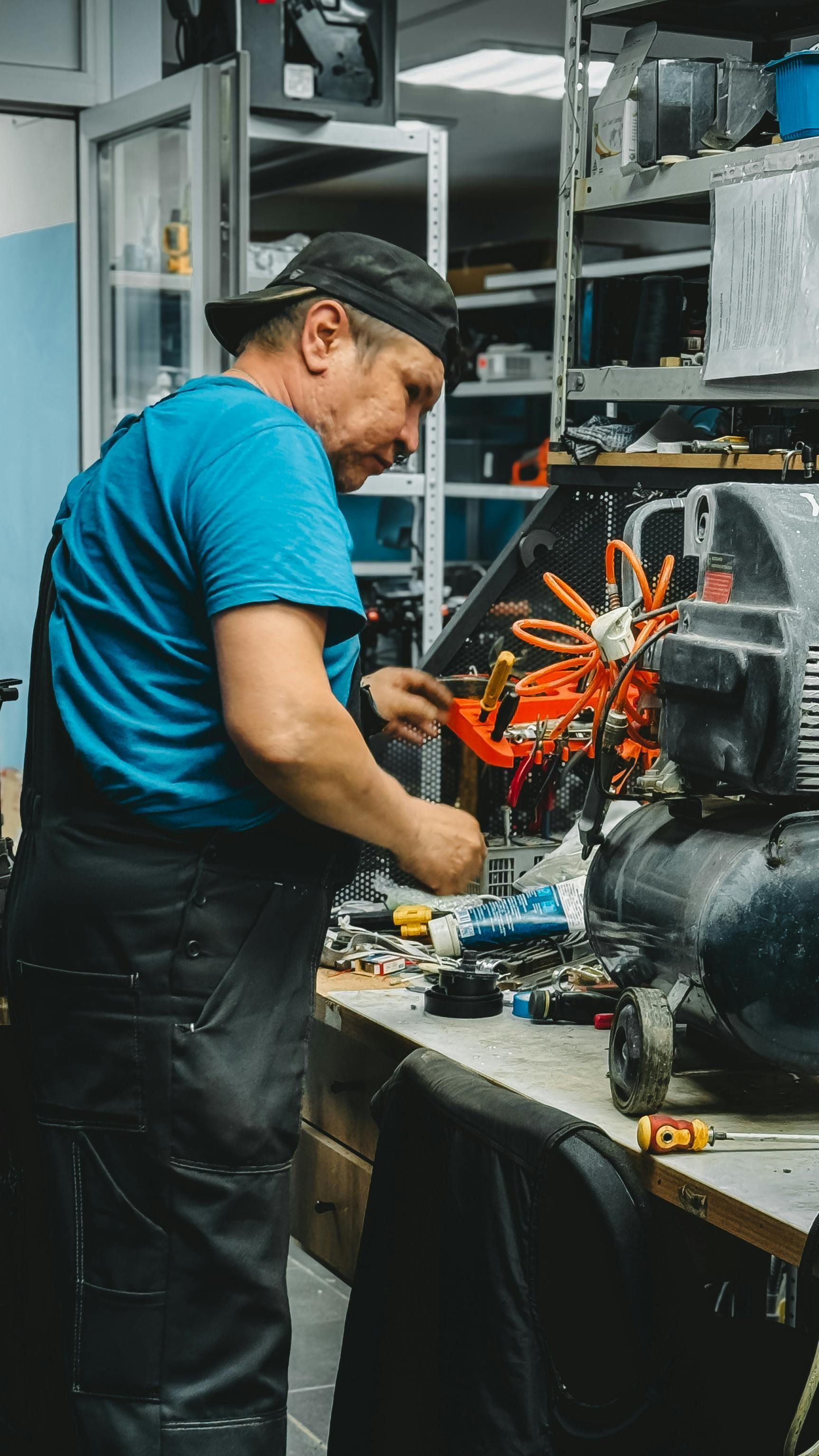Mechanic in a blue shirt working on a car engine in a workshop, with tools and shelves in the background