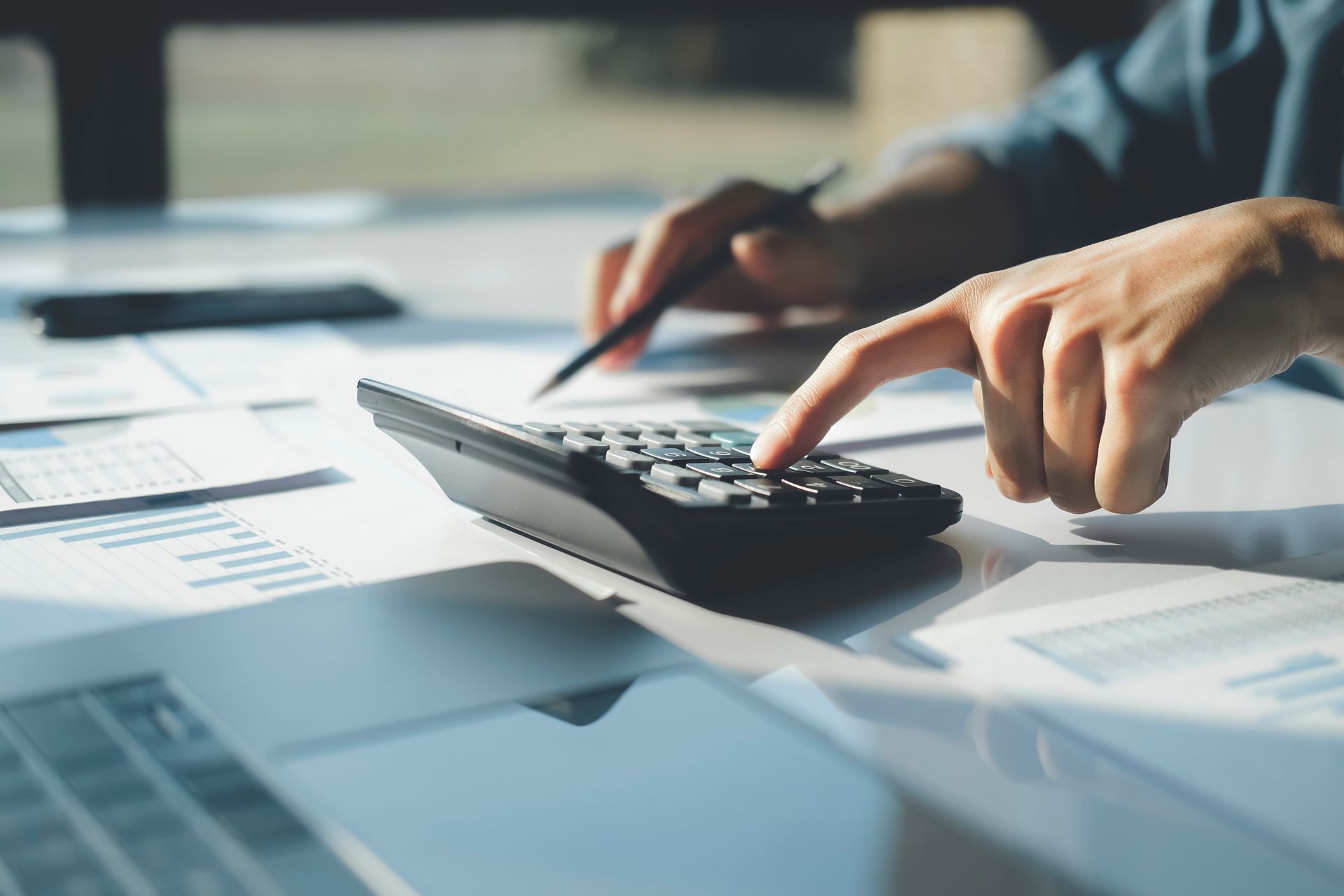 Hands using a calculator beside paperwork on a glass desk in an office setting Hands using a calculator beside paperwork on a glass desk in an office setting
