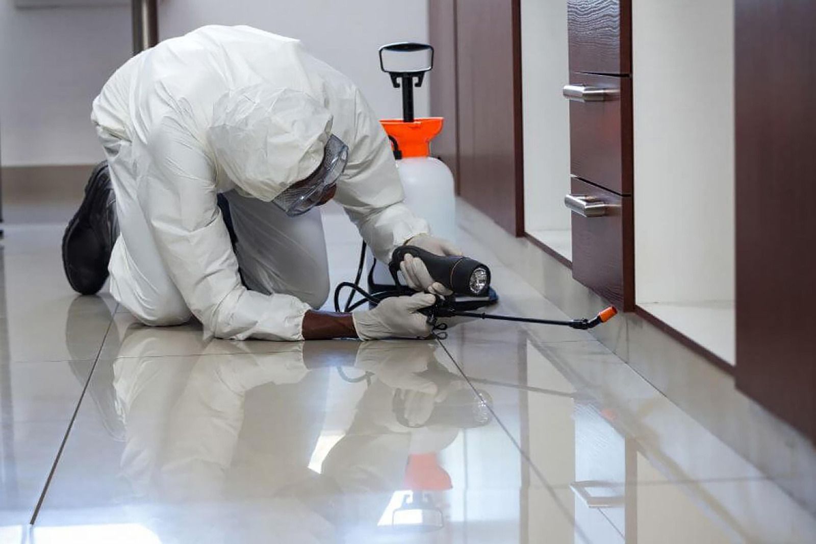 Person in protective suit spraying insecticide near a cabinet in a room.