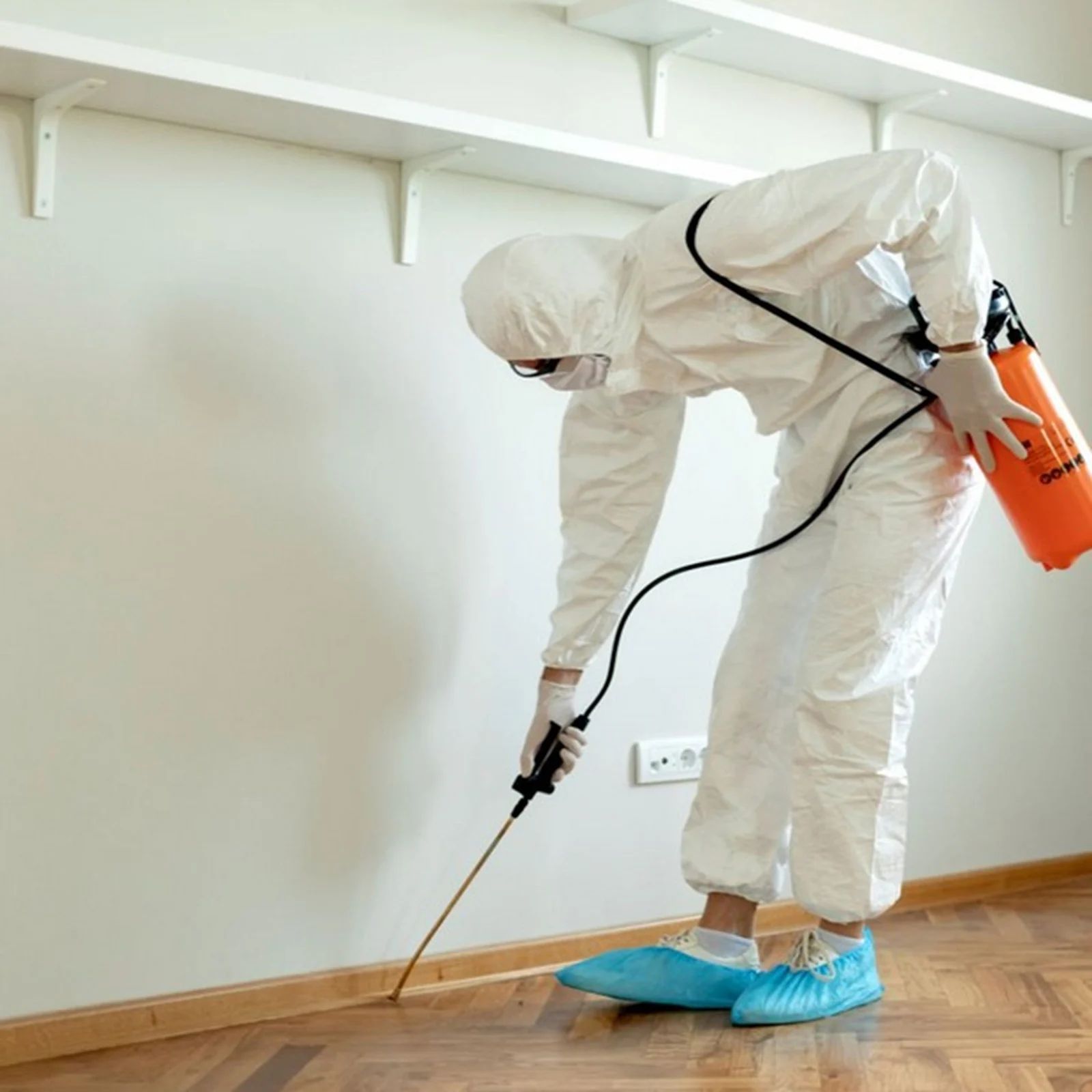 A person in protective white gear and blue shoe covers uses a chemical sprayer along the baseboard of a room.
