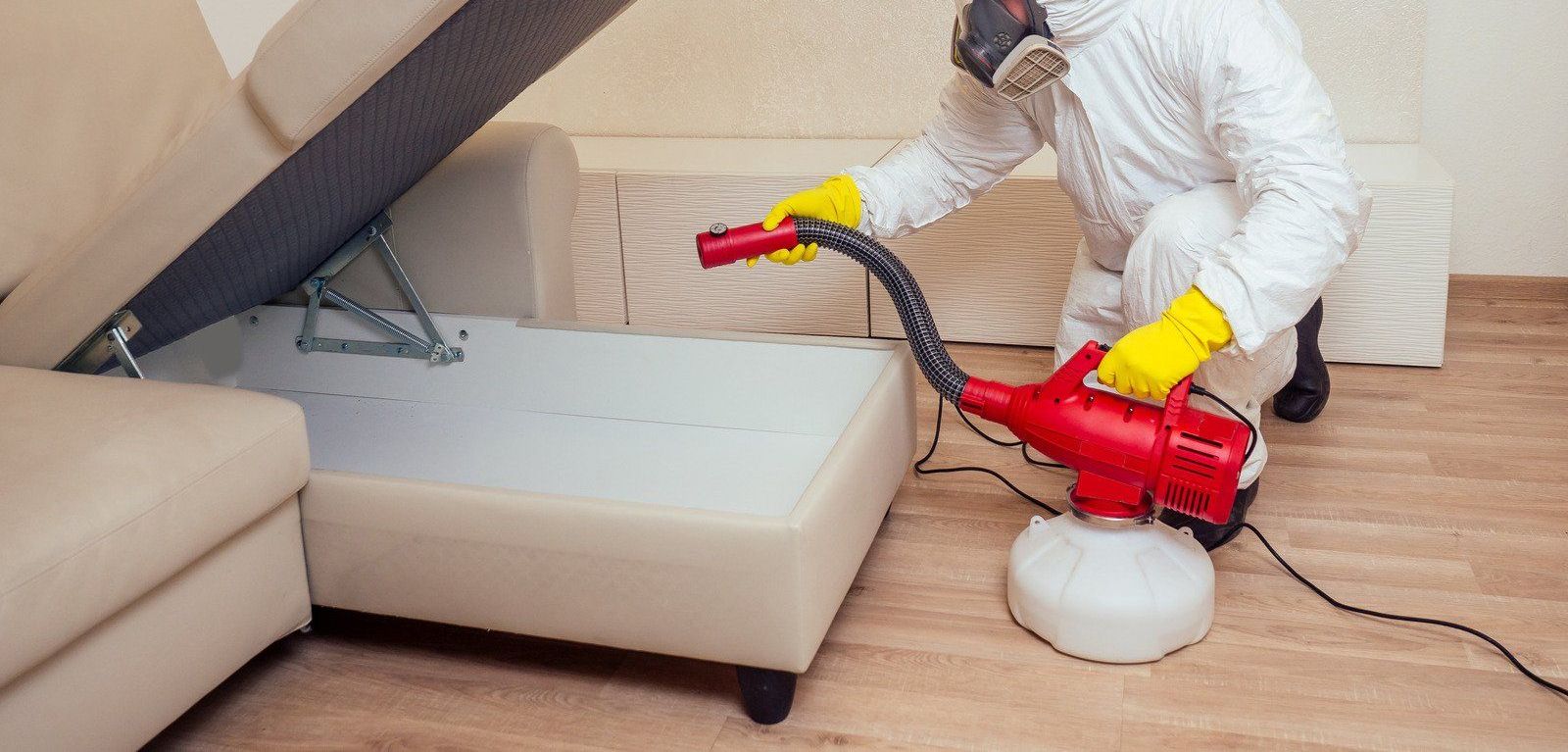 A man in a protective suit is disinfecting a couch in a living room.